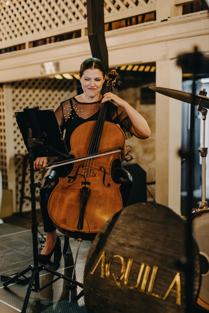 Aquila cellist performing beside the AQUILA sign
